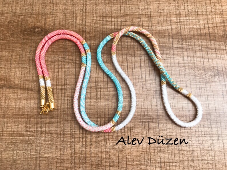May include: Two beaded necklaces, one pink and white with gold accents, and the other turquoise and white with gold accents. The necklaces are laid out on a wooden surface. Alev D&uuml;zen is written in the bottom right corner of the image.