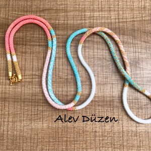 May include: Two beaded necklaces with a white, pink, and turquoise color scheme. The necklaces have gold accents and are laid out on a wooden surface. The text "Alev D&uuml;zen" is visible in the image.