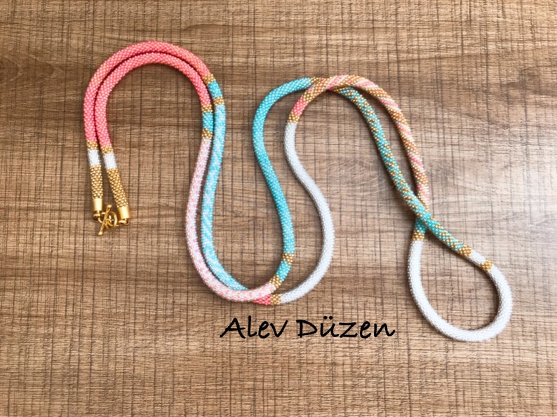 May include: Two beaded necklaces with a white, pink, and turquoise color scheme. The necklaces have gold accents and are laid out on a wooden surface. The text "Alev D&uuml;zen" is visible in the image.