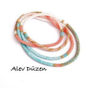 May include: A multi-strand beaded necklace with a white, peach, and turquoise color scheme. The necklace is made of small beads and has a subtle gold accent.