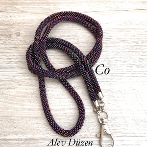 May include: A beaded lanyard with a silver clasp. The beads are iridescent and have a purple, green, and gold colour scheme. The lanyard is coiled and the text "Co" and "Alex D&uuml;zen" are visible.