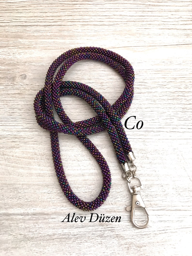 May include: A beaded lanyard with a silver clasp. The beads are iridescent and have a purple, green, and gold colour scheme. The lanyard is coiled and the text "Co" and "Alex D&uuml;zen" are visible.