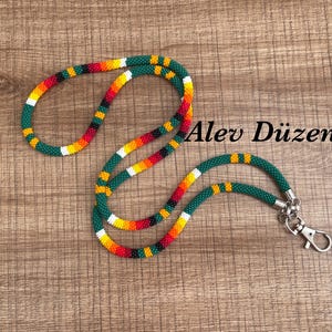 Könnte beinhalten: Ein Perlen-Lanyard mit einem sich wiederholenden Muster aus dunkelgrünen, gelben, orangen, roten, weißen und schwarzen Saatperlen. Das Lanyard verfügt über einen silberfarbenen Metallkarabiner und der Text Alev Düzen ist in Schwarz gedruckt.