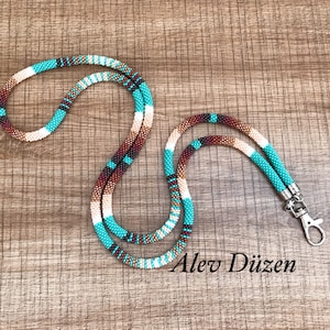 May include: A turquoise, brown and white beaded lanyard with a silver clasp. The lanyard is on a brown wooden surface. Alev Diizen is written in white text on the surface.