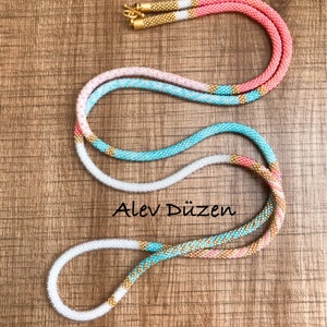 May include: A long, multi-colored beaded necklace with gold accents. The necklace is made of small, round beads in shades of white, pink, turquoise, and gold. The necklace is coiled on a wooden surface.