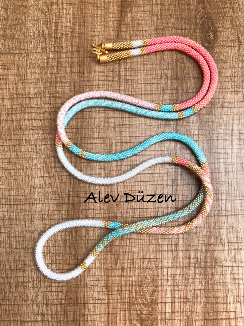 May include: A long, multi-colored beaded necklace with gold accents. The necklace is made of small, round beads in shades of white, pink, turquoise, and gold. The necklace is coiled on a wooden surface.