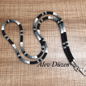 Peut inclure: Un cordon en perles noir, blanc et gris avec un fermoir argenté. Le cordon mesure environ 91 cm de long. Le texte "Alev Düzsen" est visible sur la surface en bois.