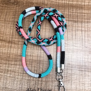 Könnte beinhalten: Ein mehrfarbiger Perlen-Lanyard mit einem silbernen Verschluss. Der Lanyard besteht aus kleinen Perlen in den Farben Rosa, Türkis, Schwarz und Weiß. Die Perlen sind in einem geometrischen Muster angeordnet.
