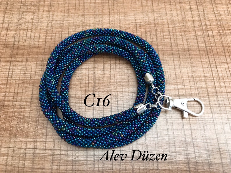 May include: A dark blue and iridescent beaded lanyard with a silver clasp and a silver chain. The lanyard is coiled and the text "Ci6" is visible in the centre of the image.