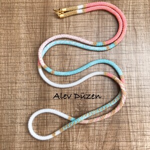 May include: A long, multi-colored beaded necklace with a gold accent. The necklace is made of white, light blue, and pink beads. The necklace is on a wooden surface.