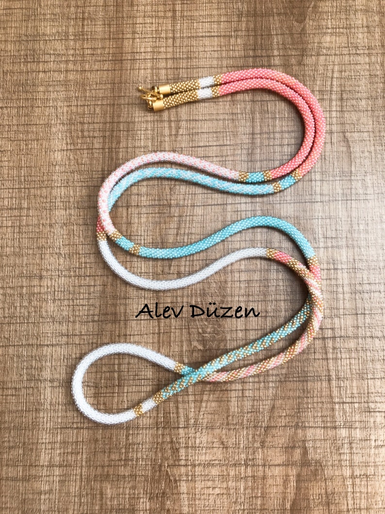 May include: A long, multi-colored beaded necklace with a gold accent. The necklace is made of white, light blue, and pink beads. The necklace is on a wooden surface.