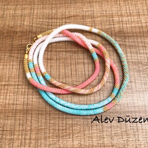 May include: Three beaded necklaces in turquoise, pink, and white with gold accents. The necklaces are layered on top of each other and are on a wooden surface. Alev D&uuml;zen is written in the bottom right corner of the image.