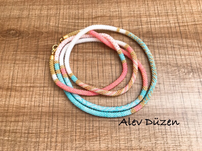 May include: Three beaded necklaces in turquoise, pink, and white with gold accents. The necklaces are layered on top of each other and are on a wooden surface. Alev D&uuml;zen is written in the bottom right corner of the image.