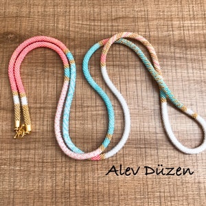 May include: Two beaded necklaces, one pink and white with gold accents, and the other turquoise and white with gold accents. The necklaces are laid out on a wooden surface. Alev D&uuml;zen is written in the bottom right corner of the image.