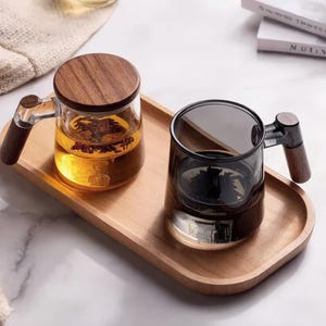 Op de afbeelding: Twee glazen theekopjes met houten handvatten en deksels op een houten dienblad. Eén kopje is gevuld met thee en heeft een houten deksel. Het andere kopje is leeg en heeft een donkergrijze tint. De dienblad is lichtbruin.
