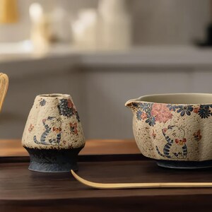 Peut inclure: Un service à thé matcha sur un plateau en bois. L'ensemble comprend un fouet en bambou, un bol à thé en céramique avec un bec verseur, un porte-bol à thé en céramique et une cuillère à thé en bambou. Les pièces en céramique sont décorées d'un chat et de motifs floraux.