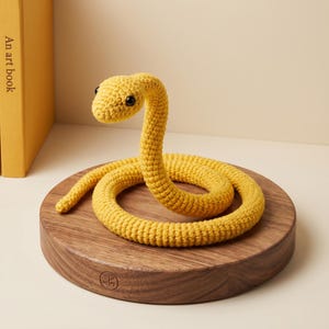 May include: A bright yellow crocheted snake toy with a textured yarn surface and two small black plastic eyes. The snake is coiled in a circular shape on a round, dark brown wooden base with a visible wood grain and a small circular logo stamp.