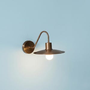 May include: A wall-mounted light fixture with a metallic bronze finish, featuring a circular base, a curved arm, and a wide, flat, disc-shaped shade. A single glowing white light bulb is visible underneath the shade, casting a soft illumination.