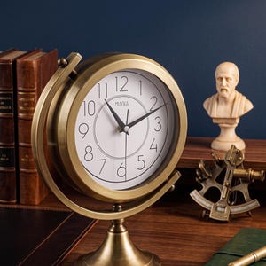 Könnte beinhalten: Eine runde Tischuhr in Messingoptik mit weißem Zifferblatt, schwarzen Ziffern und schwarzen Zeigern. Die Uhr trägt den Markennamen Muvika auf dem Zifferblatt und ist in einem kreisförmigen Kardanrahmen mit einem Standfuß montiert.