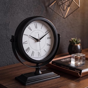 Könnte beinhalten: Eine schwarze Tischuhr aus Metall mit einem runden Rahmen und einem weißen Zifferblatt mit schwarzen römischen Ziffern und dem Schriftzug Muvika. Die Uhr ist auf einer schwenkbaren Achse an einem rechteckigen Sockel montiert und hat schwarze Zeiger.