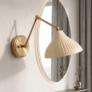 Könnte beinhalten: Eine Wandleuchte mit Schwenkarm, bestehend aus einem runden Messingsockel und einem passenden verstellbaren Arm. Die Lampe verfügt über einen breiten, cremefarbenen, geriffelten Schirm in konischer Form. Die Leuchte ist an der Wand montiert.
