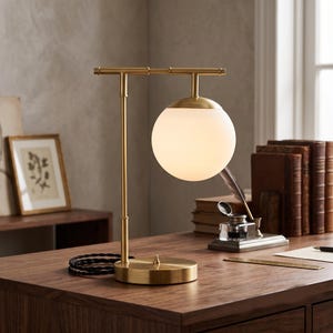 Könnte beinhalten: Eine Tischleuchte mit einem Metallrahmen in gebürsteter Messingoptik, bestehend aus einer horizontalen oberen Stange und einem runden Fuß. Ein einzelner kugelförmiger Schirm aus weißem Milchglas hängt an der Stange. Ein schwarzes Textilkabel.