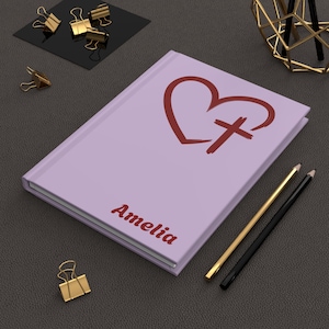 Puede incluir: Un diario lavanda con un diseño de corazón y cruz rojo, y el nombre "Amelia" impreso en la portada. También se ven lápices dorados y negros y clips dorados.