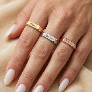 Könnte beinhalten: Drei Metallringe mit flachen, rechteckigen Oberflächen, die jeweils mit einem Namen in Schreibschrift graviert sind. Der obere Ring ist goldfarben mit dem Namen Sophia, der mittlere silberfarben mit Madison und der untere roségoldfarben mit Jennifer.