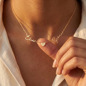 Könnte beinhalten: Eine goldfarbene Halskette mit einer feinen Kette, die zwei in Schreibschrift geschriebene Namen, Elia und Giana, zeigt, welche durch ein kleines, mittiges Herz verbunden sind. Das Metall ist poliert und die Schrift ist in die Kette integriert.