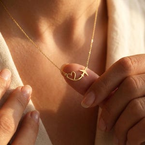 May include: A gold-toned necklace featuring a delicate chain and a pendant shaped like a small airplane flying toward a heart outline. The pendant is held by fingers, showcasing the metallic finish and the connected design of the charm.