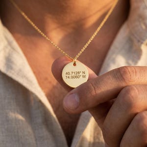 May include: A gold-toned circular pendant necklace featuring the engraved coordinates 40.7128° N 74.0060° W. The pendant is suspended from a delicate gold-toned chain, showcasing a polished metallic finish and clear, dark, centered text engraving.