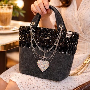 May include: A dark gray denim handbag featuring a black textured upper section, silver chain accents, and a heart-shaped charm covered in multicolored crystals. A small white tag with the word coffee is attached to the side of the bag.