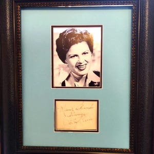 May include: A framed black and white portrait of a woman with dark hair, above a handwritten signature. The portrait is set against a light blue background within a dark, ornate frame.