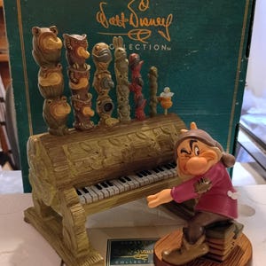 Könnte beinhalten: Eine Walt Disney Classics Collection Figur, die Grumpy zeigt, wie er auf einem Stapel Bücher sitzt und ein baumstammförmiges Klavier spielt. Das Klavier hat schwarz-weiße Tasten und ist mit sechs geschnitzten Tiertotems und einem kleinen Vogel verziert.