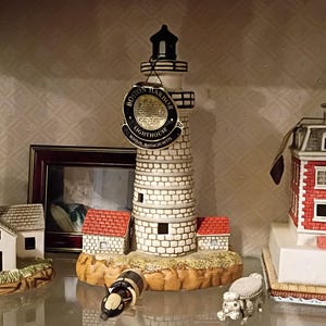 May include: Three decorative lighthouse figurines. The left one is white with a red stripe. The centre one is a stone-textured Boston Harbor lighthouse with a circular tag. The right one is a red brick building with a white base and a red roof.
