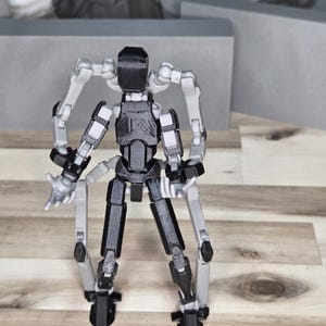 May include: A black and gray articulated robot figure with multiple limbs and a rectangular head. The robot has a detailed design with visible joints and is posed on a wooden surface.