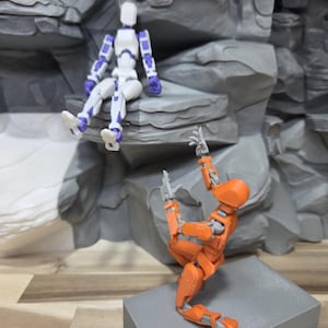 May include: Two articulated action figures, one white and purple, the other orange, are posed against a gray rock formation. The white figure is seated, while the orange figure appears to be climbing a gray block.