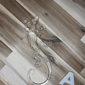 May include: A clear, decorative lizard figurine with intricate detailing. The lizard is positioned on a wooden surface, with its tail curled. A white letter "A" is also present.