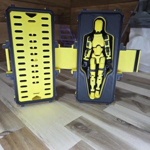 May include: A yellow and black action figure inside a black case with yellow hinges. The case is open, revealing the figure in a molded interior. The figure is yellow with black accents, and the case is on a wooden surface.