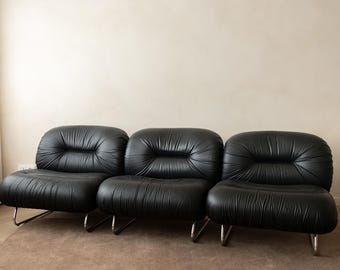 May include: A three-seat sofa upholstered in black leather with a tufted, rounded design. The sofa features three distinct, plush sections with deep indentations in the center of each backrest and seat, supported by thin, curved metal legs.