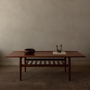 Puede incluir: Una mesa de centro de madera de estilo moderno de mediados de siglo, con un estante inferior y una parte superior rectangular. Un cuenco de madera oscura y un objeto decorativo de metal están sobre la mesa. La mesa está sobre una alfombra marrón.