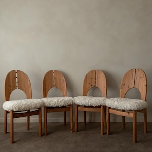 Puede incluir: Un conjunto de cuatro sillas de madera con marcos de grano de madera marr&oacute;n claro y respaldos redondeados con un corte vertical central. Cada silla tiene un coj&iacute;n de asiento grueso de piel sint&eacute;tica blanca texturizada sobre cuatro patas.
