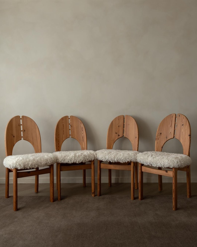 Puede incluir: Un conjunto de cuatro sillas de madera con marcos de grano de madera marr&oacute;n claro y respaldos redondeados con un corte vertical central. Cada silla tiene un coj&iacute;n de asiento grueso de piel sint&eacute;tica blanca texturizada sobre cuatro patas.