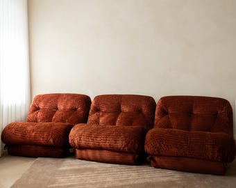 May include: A three-piece modular sofa set upholstered in a deep rust-colored, textured velvet fabric. Each individual seat section features a low-profile design with tufted backrests and thick, plush cushions, creating a continuous seating arrangement.