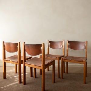 Puede incluir: Un conjunto de cuatro sillas con marcos de madera de color marrón claro y asientos y respaldos de cuero color canela. Cada silla tiene una base de patas rectas y un panel de respaldo de cuero curvado unido al marco de madera.