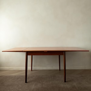 Puede incluir: Una mesa de comedor rectangular de madera con un acabado marrón cálido y una superficie lisa. La mesa cuenta con cuatro patas cónicas y dos hojas laterales extensibles que están completamente desplegadas, creando una superficie amplia y plana.