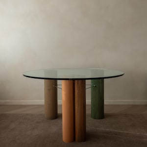 Puede incluir: Mesa redonda con tapa de cristal y tres patas cilíndricas. Las patas están pintadas en tonos beige, naranja y verde. La mesa está sobre una alfombra marrón contra una pared neutra. Diámetro aproximado 100 cm.