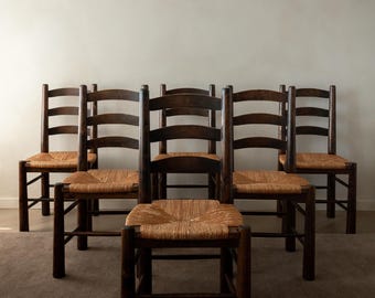 May include: Six dark wooden chairs with woven straw seats. The chairs have a ladder-back design and are arranged in a semi-circle. The chairs are set against a neutral-colored wall and carpeted floor.