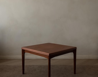 May include: A square, brown wooden table with four straight legs. The table is made of a dark wood and sits on a brown carpet. The background is a neutral, light-colored wall.