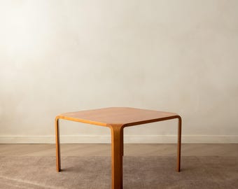 May include: A square wooden coffee table with a smooth, light brown finish. The table features four legs that curve seamlessly from the tabletop, creating a continuous, rounded silhouette with soft edges and a flat, square surface.